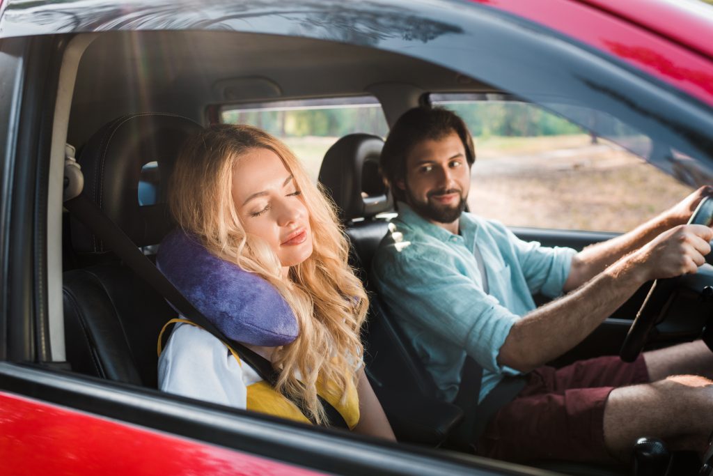 Woman falls asleep in the passenger seat of a car with a travel pillow round her neck