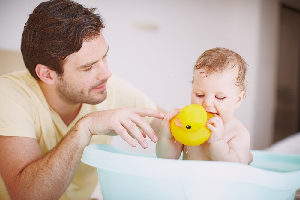 Dad plays with his baby who's in the bath chewing a yellow rubber duck