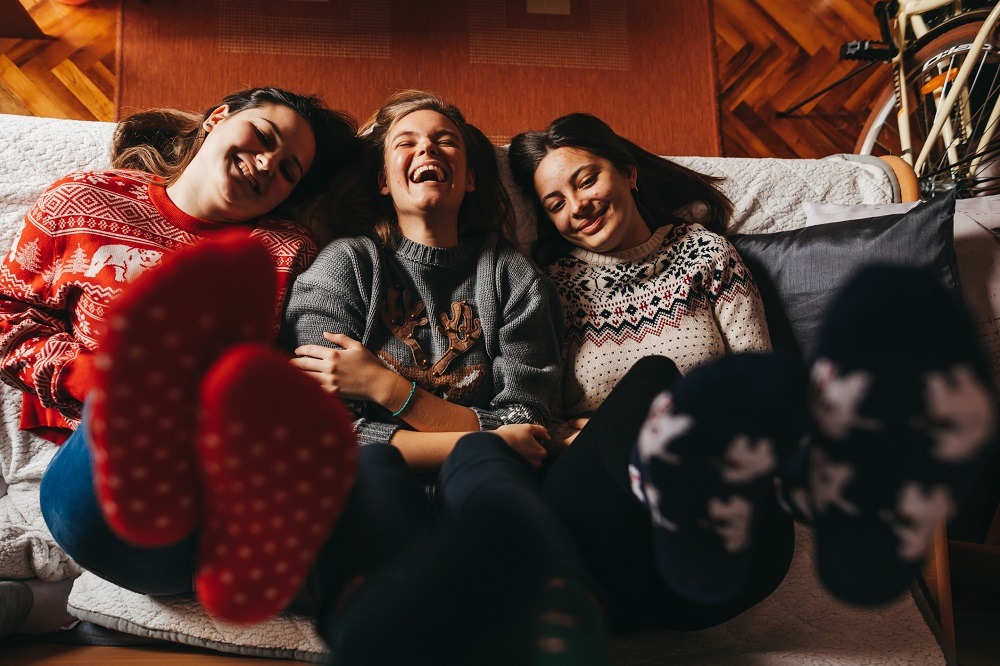 Three young female friends dressed in winter clothes sit on the sofa laughing together