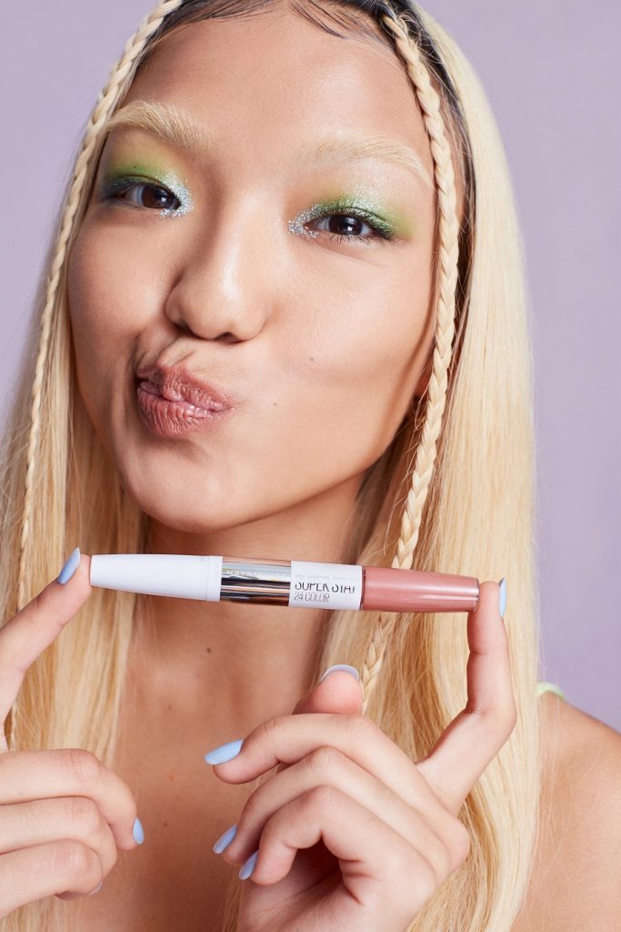 Girl with blonde hair and green eyeshadow poses with a Maybelline lipgloss