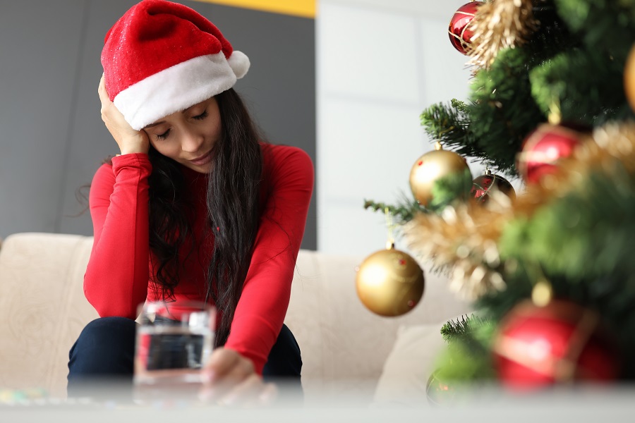 Woman wearing a red santa hat holds her head as she nurses a hangover next to a Christmas tree and glass of water