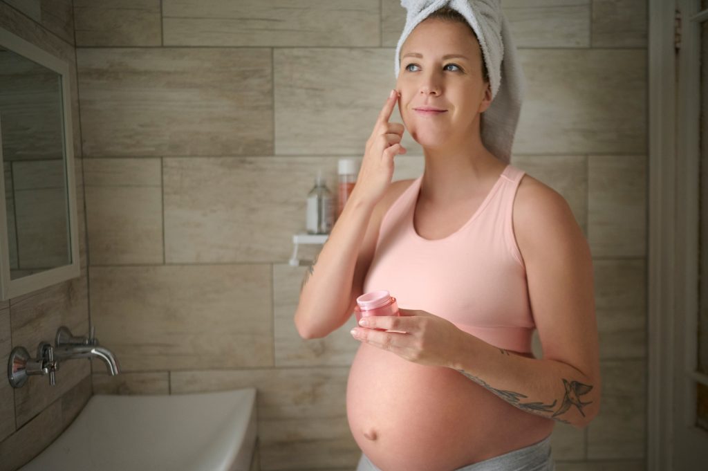 Pregnant woman smiling in bathroom applying moisturiser to face